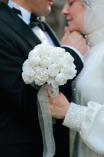 A bride and groom are captured during a wedding ceremony, with the bride holding a bouquet of white roses and peonies wrapped in sheer white ribbon. The groom is dressed in a black tuxedo with a white