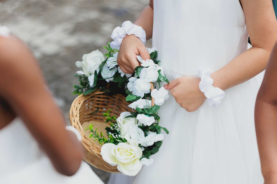 A close-up of a person holding a floral arrangement consisting of white roses, peonies, and smaller white flowers with green foliage, arranged in a curved pattern. The bouquet is framed by a wicker ba