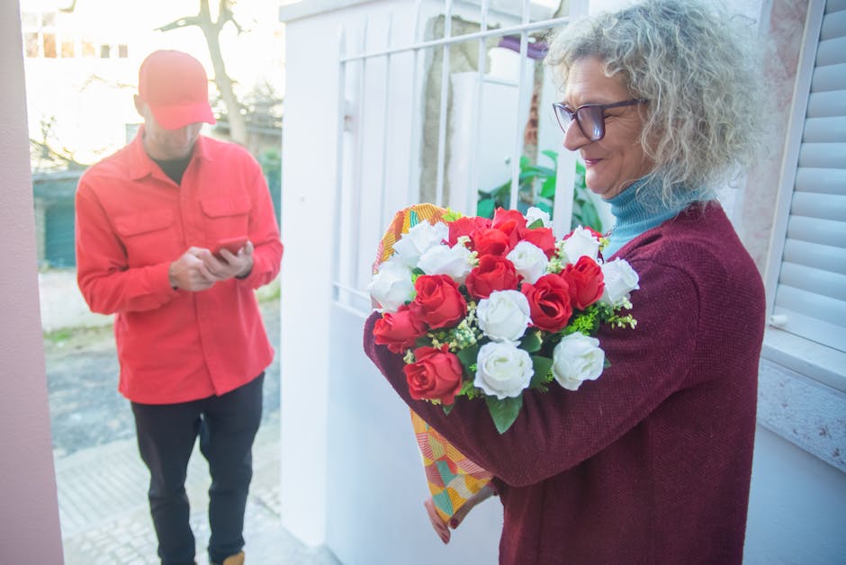 A woman with curly gray hair and glasses, dressed in a burgundy sweater and light blue turtleneck, holds a bouquet of fresh red and white roses wrapped in colorful patterned paper. She stands near a w